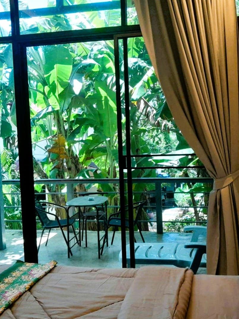 Modern Bungalow 1 – vom Bett durch die Balkontüren: Tisch und Stühle, eine Liege und die Bananenstauden hinter dem Betonbalkon