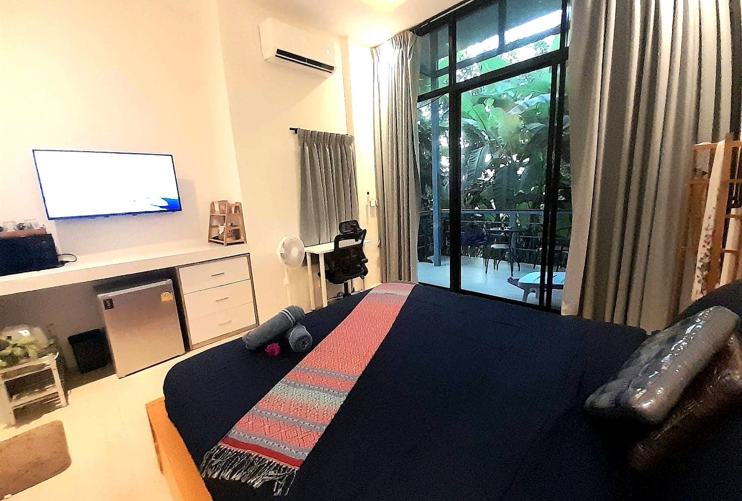 Bungalow interior today: desk with a monitor, fridge, air conditioning and floor-to-ceiling balcony doors onto the jungle